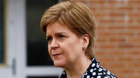 Nicola Sturgeon - woman with short red hair, wearing a black blouse with white floral detailing and looking to the left side of the frame