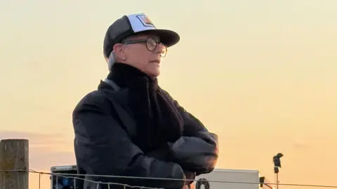 BBC Jamie Lee Curtis behind a fence, wearing black jacket and cap