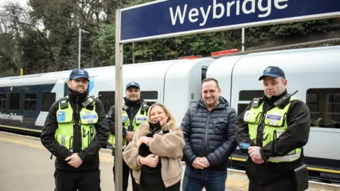 Tilly, a black cat, with her owners and three members of rail staff. They are stood on the platform. 