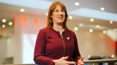 PA Media Rachel Reeves speaking to someone out of shot, gesturing with her hands. She is wearing a red top and a yellow poppy.