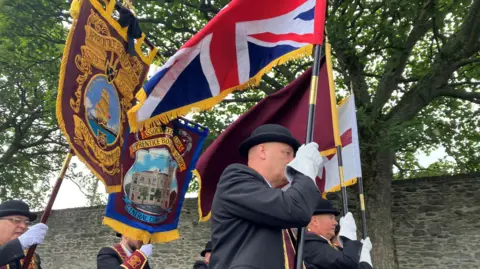 A group of men wearing black suits and black bowler hats marching in a parade. They are wearing maroon sashes an carrying various flags. There is a band behind them and old walls to their left.