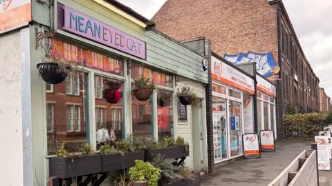 BBC / Bethany White The outside of the Mean Eyed Cat, which is a small pub with a green exterior and pink sign. There are hanging baskets outside the shop, with large planters running along the windows. There is a sports shop next door and a row of terraced brick houses to the right.