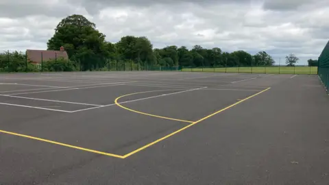 Two tarmac tennis courts that are fenced in with green fencing. There is a house in the distance and trees and fields. 