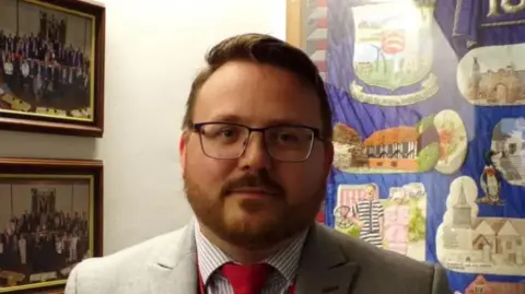 Daniel Cowan wearing a light grey blazer and a red tie. He is wearing a blue striped shirt. He has glasses and brown hair with a beard. Behind him on a wall are some photographs showing groups of people.