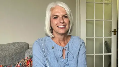Sarah Thyer sitting on her grey sofa at her home in Swansea. She is wearing a pale blue shirt with an embroided floral design with a string tie affixing it at the top. Sarah has shoulder length straight white hair and she is smiling. Behind her is a glass-panelled living room door and the top of a multi-coloured pillow can be seen over her right shoulder.