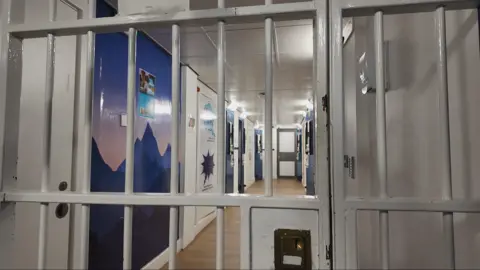 BBC A photo of the bars of a police custody cell. Through the bars is a corridor which has pained artwork on the walls and messages