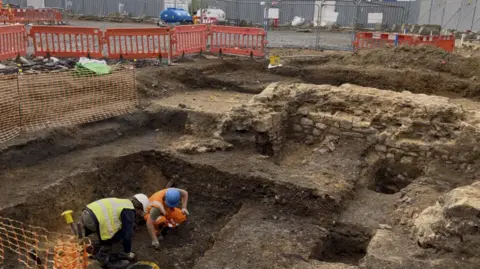 Archaeologists dig in a pit near a stone wall 