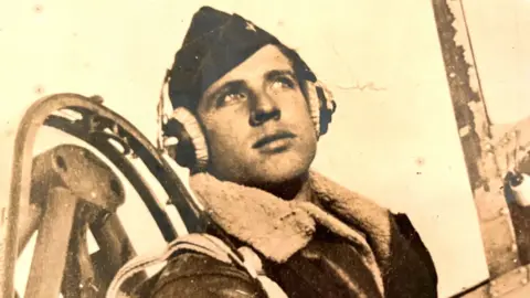 An old sepia photograph. It shows a young man wearing a leather flight jacket with a sheepskin collar. He is also wearing an air force cap and a pair of headphones over his ears. What looks like part of a WW11 airplane cockpit is behind him  