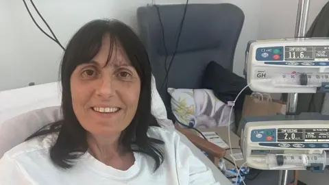 Nicola smiling at the camera. She is in a hospital bed with a monitor beside her. There is a grey hospital chair to the right which has a cushion and some parcels on it.