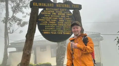 Freddie wears an orange raincoat and points to a sign that reads Kilimanjaro national park, that also includes timings to different points within the park.  It is very misty and wet.