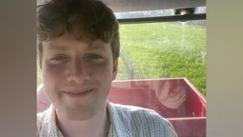 Jon Watt sitting in the cab of a tractor. He has mid-brown short hair, curling on top, and is wearing a checked shirt. Behind him through the rear window is a red trailer and beyond that there are fields. 