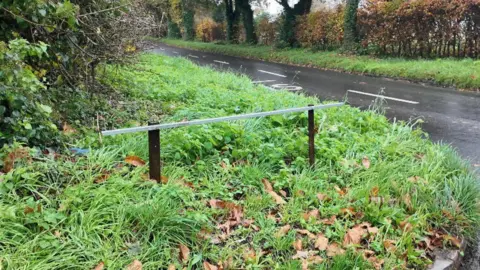 BBC A road sign on a grass verge which has been stolen. All that is left is two small metal posts and a metal bar where the sign was once attached.