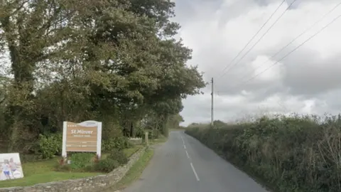 A view of a country road. A St Minver Holiday Park sign is on the left hand side of the road. Trees and bushes line either side of the road.