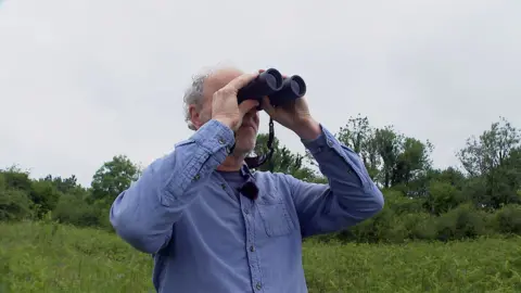 Charlie Elder is pictured though his face is not really visible as he is holding black binoculars to his eyes. He is wearing a blue shirt over a blue T-shirt and he has curly grey hair.