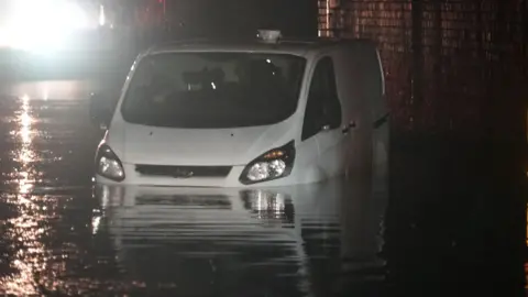 Van stuck under railway bridge on A429 near Hullavington. It is dark and the water level is up to just below the headlights. The van seems to have been abandoned. 