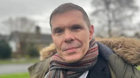 Michael is wearing a dark coat with a fur-lined hood and a multicoloured striped scarf wrapped around the neck. Underneath the coat, a white shirt and a green patterned tie are visible. The background shows a grassy area with a road, trees without leaves, and houses in the distance under an overcast sky.