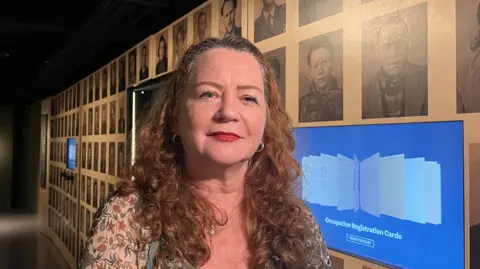 A woman with long brown curly hair standing in front of a wall with a number of portrait black and white photos on it