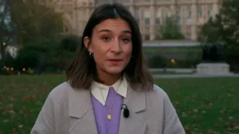 A woman with brown hair, wearing a grey jacket and purple jumper
