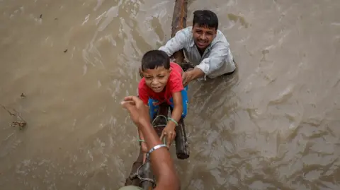 Reuters Seorang pria membantu putranya memanjat jalan layang yang sedang dibangun setelah mengungsi akibat naiknya permukaan air sungai Yamuna setelah hujan lebat di New Delhi, India. Laki-laki itu berada di air berlumpur sampai pinggang.