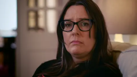 Firecrest Films Head shot of a woman with long black hair, photographed while seated in a living room with sad expression on her face.