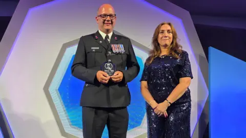 Martis Media PC Jay Wakefield, a tall bald man wearing glasses and dark grey uniform decorated with military medals, collects his award from the Police Federation's national deputy secretary Mel Warnes, a woman with long brown hair, wearing a long sparkly navy blue dress