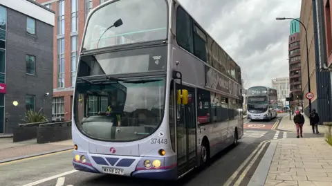 Oli Constable/BBC A First Bus stopped on a city centre road. The bus is coloured various shades of purple with some advertisement on its sides. People walk on the pavement next to it.