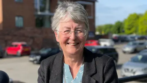 A woman with short, greying hair and rimless glasses smiling at the camera. She is standing in front of a car park and red-bricked building wearing a dark blazer over a blue blouse. 