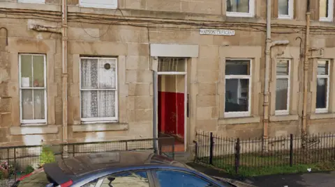 Google Exterior of a flat. A sign above the door says Watson Crescent. There are railings outside and the paint inside the doorway is red.