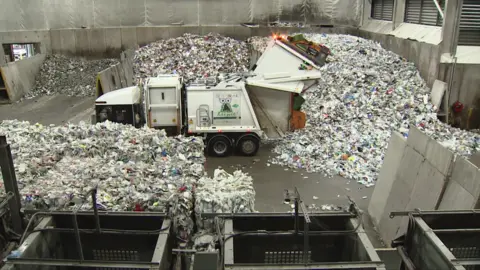 Inside a large waste‑processing facility, a rubbish truck is tipping a load of mixed recyclable materials onto a massive pile. The truck is surrounded by heaps of plastic, paper, and other waste, with concrete walls and industrial equipment visible around the sorting area.