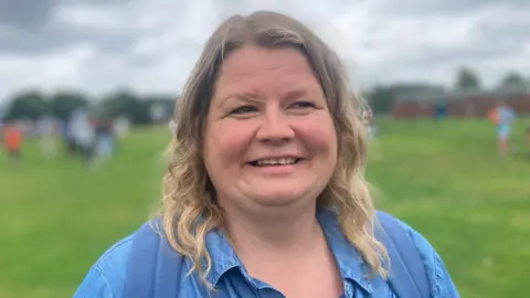 A woman with shoulder-length blonde hair smiles and looks to the right of the camera. She is wearing a blue top and a backpack. Behind her is a large area of grass and, out of focus, a crowd of people.