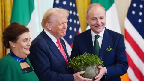 EPA Three people stand smiling. On the left, a woman with brown hair wearing a green dress. In the middle, a man with blonde hair wearing a navy suit with a red tie. On the right, a balding man in a navy suit and dark green tie. The man on the right has a sprig of shamrock in his breast pocket. The two men are holding a bowl - inside the bowl is a green plant. 