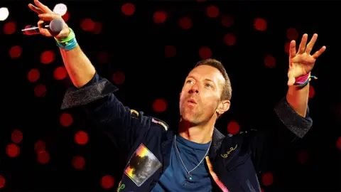 Getty Images Coldplay's Chris Martin holds both hands in the air, with a black microphone in his right hand. he wears a denim jacket and a blue top underneath, with red spotlights behind him.