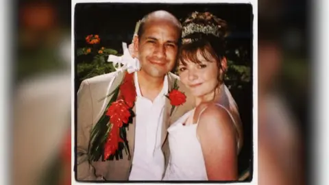 Nicky Wake A couple's wedding picture. The person on the left, Andy, is a bald, tanned man wearing a beige jacket and white shirt with a red flower garland around his shoulders. Nicky stands next to him wearing a white dress with her hair in an updo. 