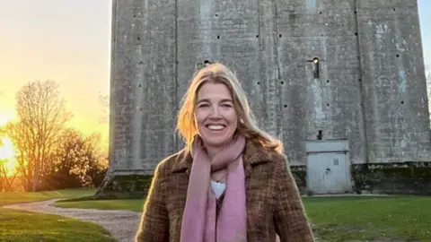 A head and shoulders image of Demetra Lindsay, co-owner of Hedingham Castle. She is wearing a pink scarf and a coat over a white top. She is smiling and standing in front of Hedingham Castle. 
