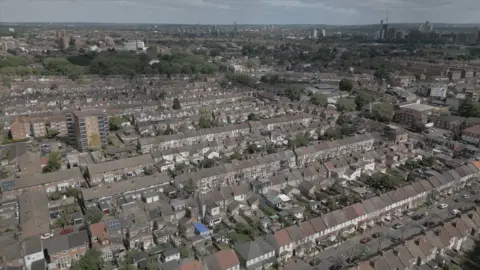 A still of an aerial drone shot showing houses near the Greenway in east London. Roofs of mainly terraced houses can be seen with trees in the distance.