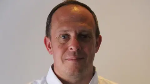Chris Gnapp is a man with brown short hair, blue eyes and a white shirt . He looks at the camera and is wearing a white shirt. He is standing in front of a cream wall. 