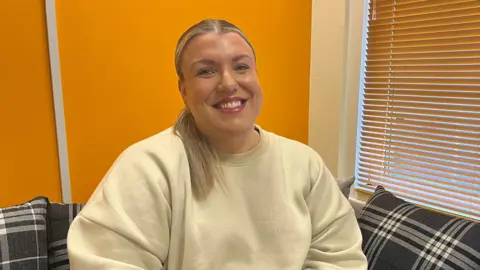 Bianca Nelson wears a cream sweatshirt. She is sitting on a sofa which has a white and grey check pattern. She is smiling warmly at the camera and wears he long blonde hair tied back in a ponytail. 