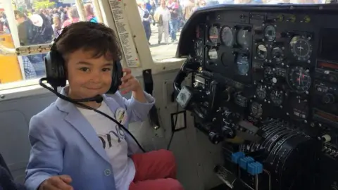 Family handout A very young boy in the cockpit of a plane. He has headphones on which have a mic from the headset. They are too big for his head. The boy is smiling at the camera. You can see the switches and dials of the cockpit.
