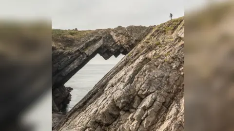 Quintin Lake A jagged rocky outcrop with a small figure of a man on top of the rocks. The rock has a bridge-like formation, with the sea visible through a hole under the rock connecting two points.