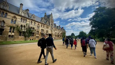 People walking in front of Christ Church Meadow in Oxford. Everyone is walking away from the camera. It is a cloudy day.