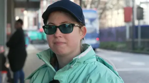 Rebecca Clarke is seen wearing sunglasses, a dark cap and a light green jacket at a bus stop, with a bus and other passengers visible in the background.