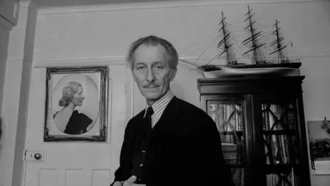 Getty Images A black and white photo of a man, wearing a cardigan and a shirt and tie, standing inside a room. There is a bookcase behind him and a painting of a woman hanging on the door. 