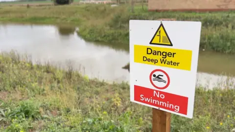 A sign reading "Danger Deep Water" and "No Swimming" is fixed to the top of a wooden post. It stands on vegetated land in front of a small body of water, with the land on the other side also visible.