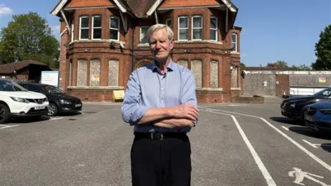 A man with grey hair in a blue shirt with the sleeves rolled up and black trousers standing in front of a boarded up building and a number of cars.