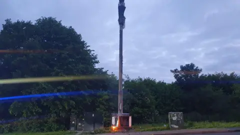A 5G mast is on fire. Trees and bushed are in the background. It's late evening.