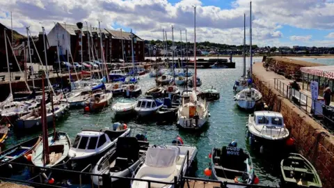Graham Christie A colourful harbour filled with sailboats and small motorboats moored along stone quays. Bright blue skies and large white clouds overhead add contrast to the bustling waterfront and the buildings lining the harbour.