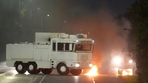 A white water cannon vehicle next to flames on the road
