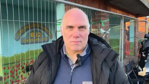 A man with a thick black coat and blue fleece looks at the camera standing outside a bus shelter. 