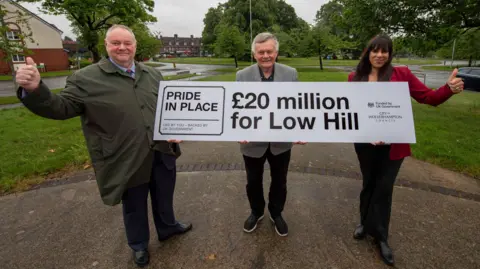 City of Wolverhampton Council A man wearing a coat, shirt and tie is on the left, another man with grey hair is wearing a grey jacket in the middle and a woman is on the right in a red jacket. They are carrying a large banner including "£20 million for Low Hill" in black letters and numbers.
