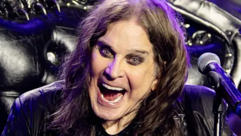 Ozzy Osbourne on stage with his arms outstretched and a big open-mouthed grin